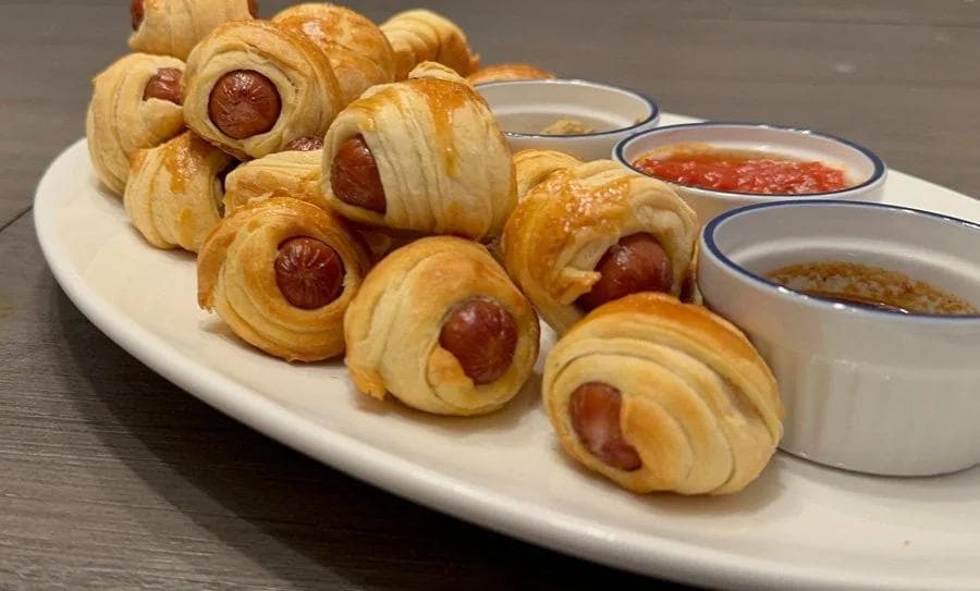Air fryer pigs in a blanket on a white plate with three small bowls of dipping sauces.