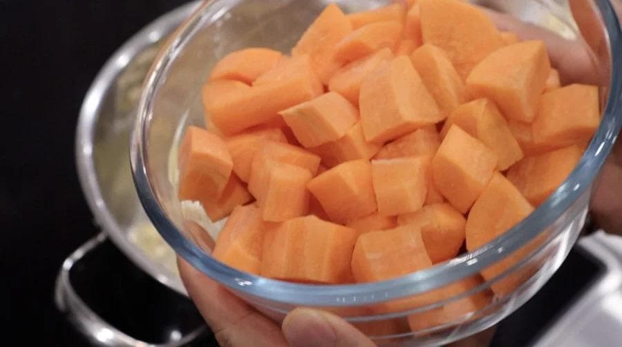 Diced carrots in a glass bowl being held over a pot during soup preparation.