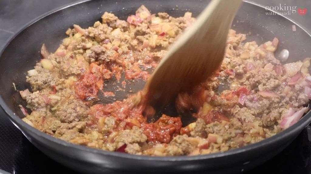 Wooden spoon stirring cooked ground beef with tomato paste in a dark skillet during beef martabak preparation.