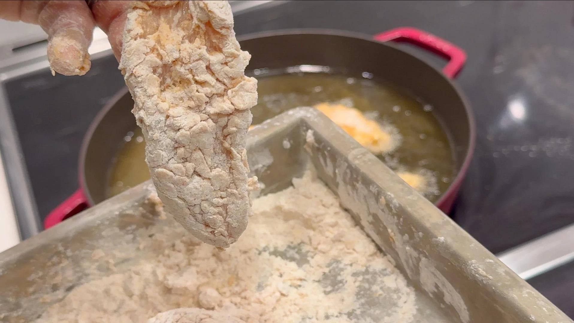 Chicken tender being coated in seasoned flour mixture over a shallow pan with oil heating in the background.