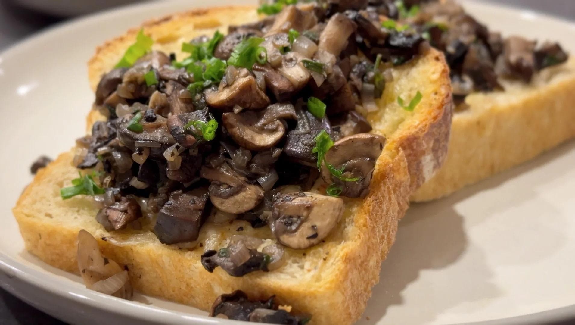 Sautéed mixed mushrooms served over toasted bread, garnished with fresh green onions on a white plate.