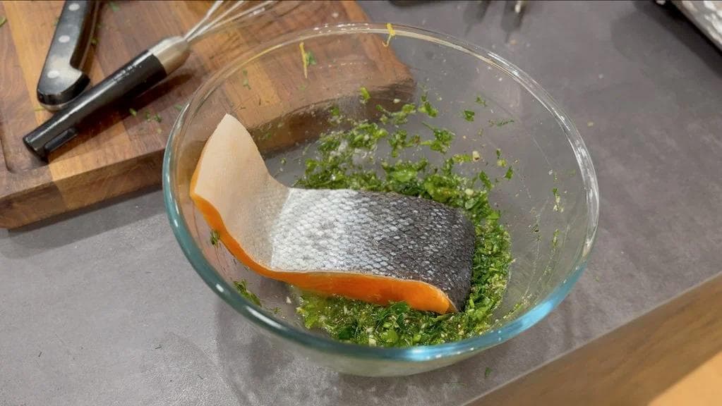 Raw salmon fillet coated with herb marinade in a glass bowl, surrounded by chopped parsley and dill.