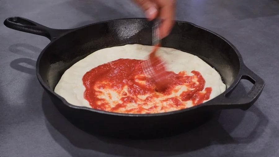 Red pizza sauce being spread onto white dough in a cast iron skillet.