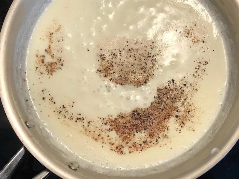 Creamy béchamel sauce in a pan with nutmeg and black pepper seasoning sprinkled on top.