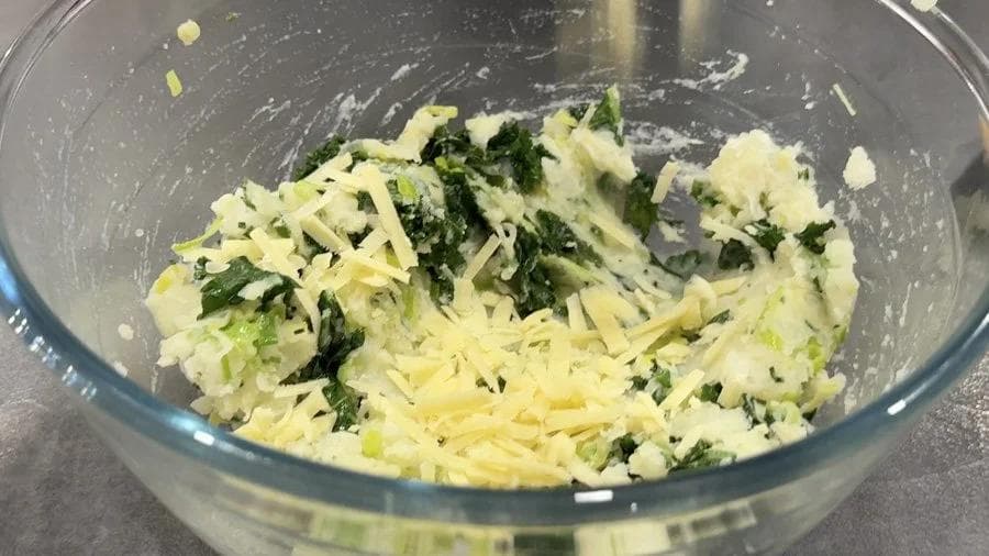 Mashed potatoes mixed with shredded cheddar cheese and chopped green onions and chives in a glass bowl.