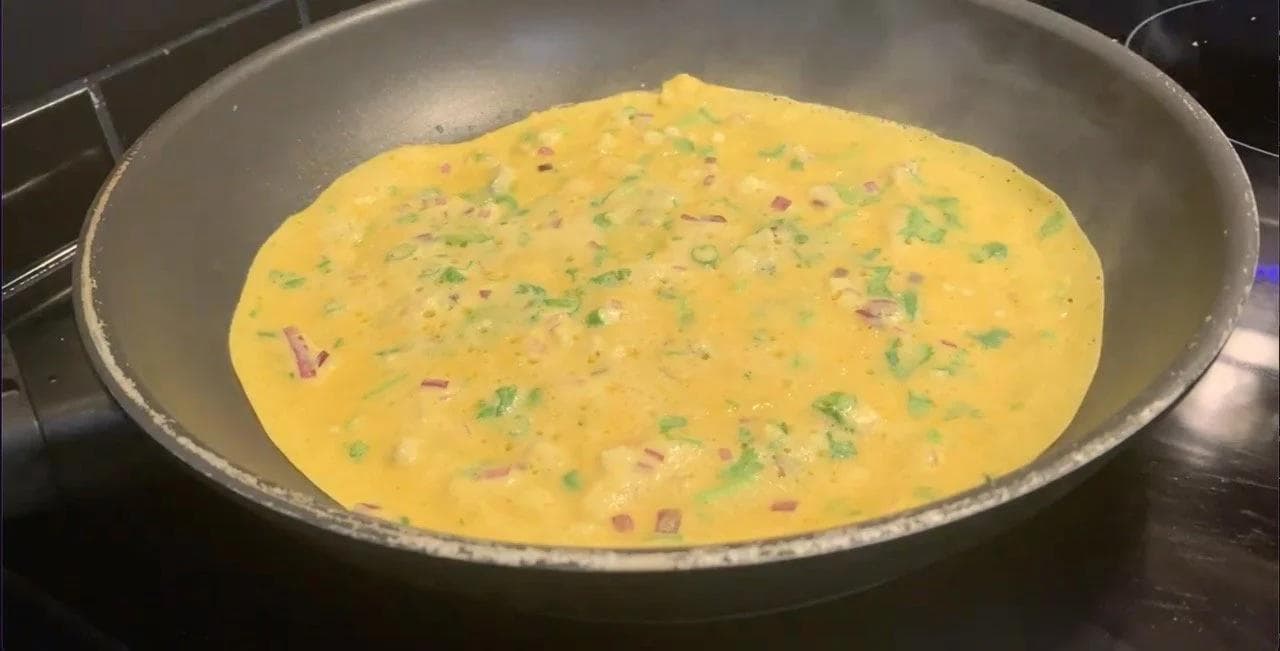 Golden besan chilla cooking in a pan, topped with cilantro and red onions, edges beginning to brown.