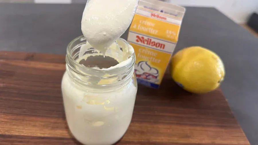 Homemade mascarpone cheese being spooned into a mason jar for storage, with butter and lemon nearby.