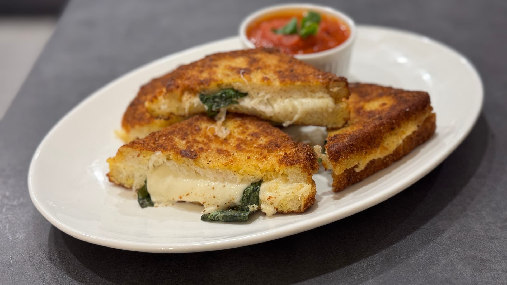 Golden-brown mozzarella in carrozza sandwiches with melted cheese and basil, served with red sauce on white plate.