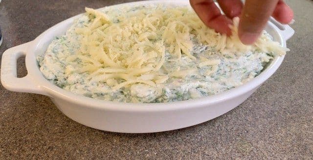 Creamy spinach dip mixture in a white casserole dish with melted mozzarella cheese being stretched with a hand.