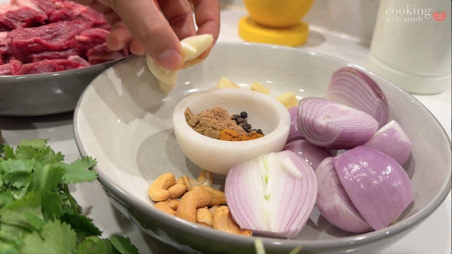 White bowl with sliced red onions, cashews, spice paste in small mortar, cilantro, and hand adding potato slice above.
