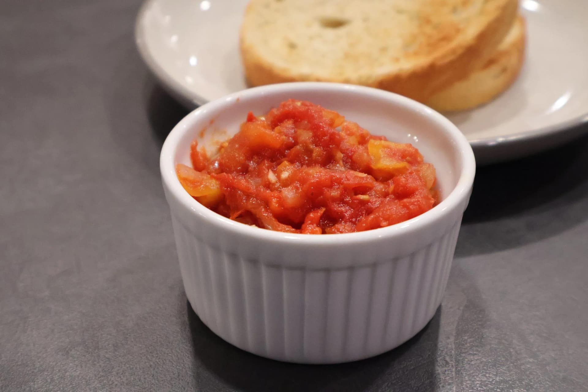 White ramekin filled with chunky red Piperade sauce, served alongside toasted bread slices on a plate.