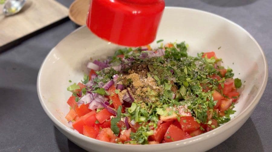 Pico de gallo ingredients mixed in white bowl: diced tomatoes, red onions, cilantro, and spices with red bottle in background.