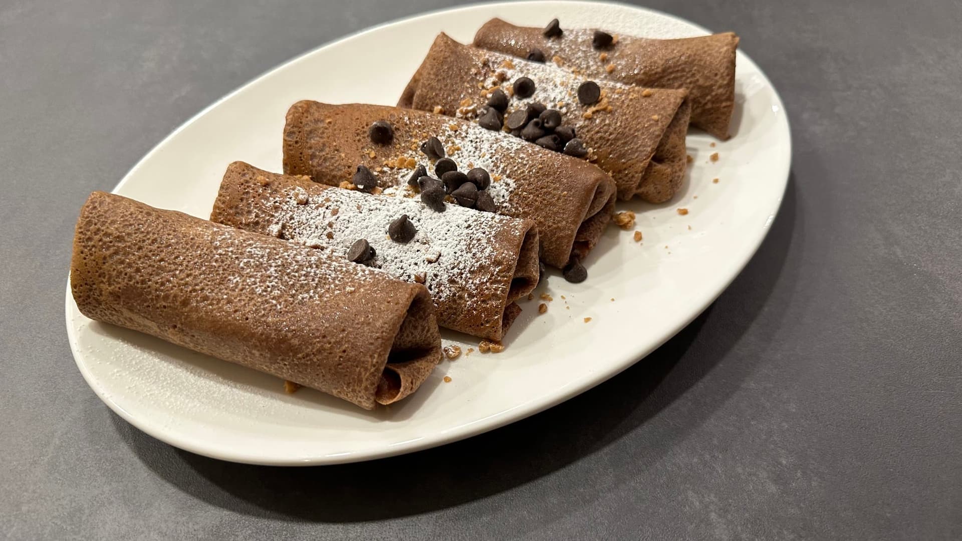 Chocolate crepes dusted with powdered sugar and topped with chocolate chips on a white plate.