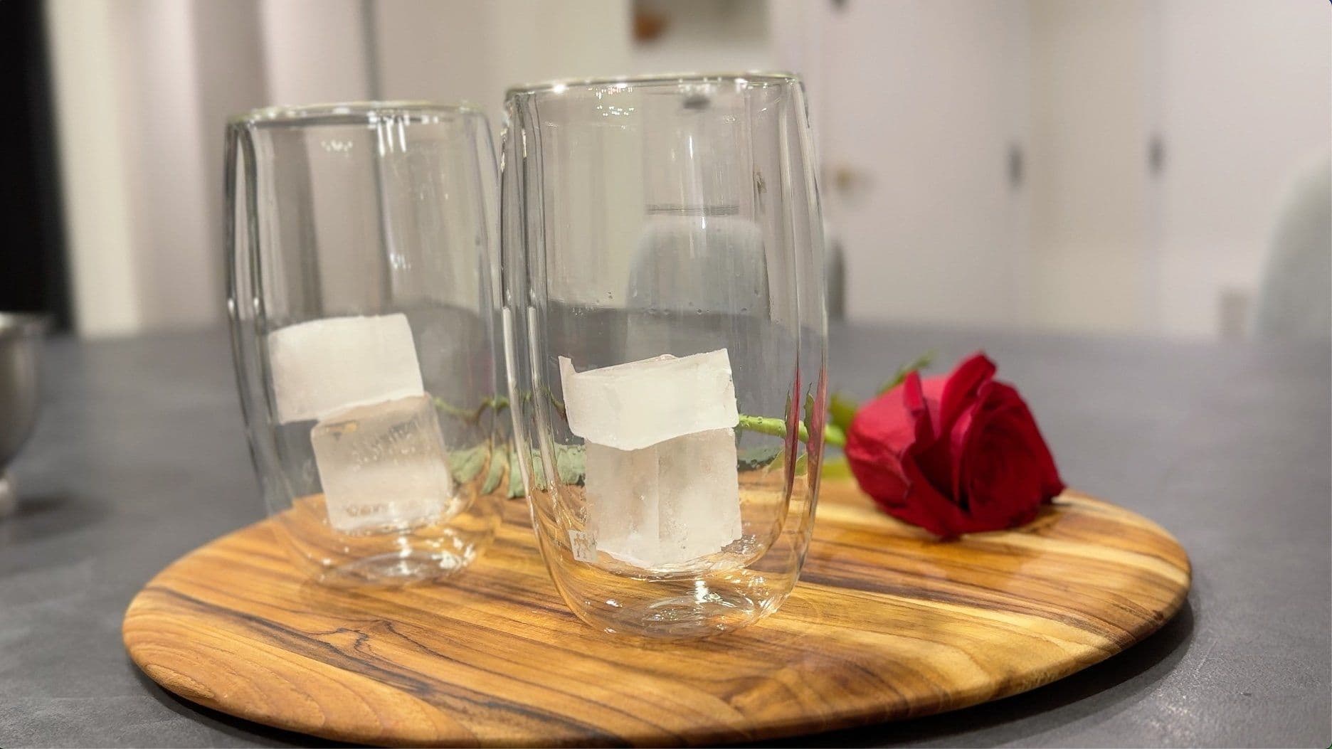 Saffron Pista Lassi served in tall glasses with ice cubes on a wooden board, garnished with pistachios and a red flower.