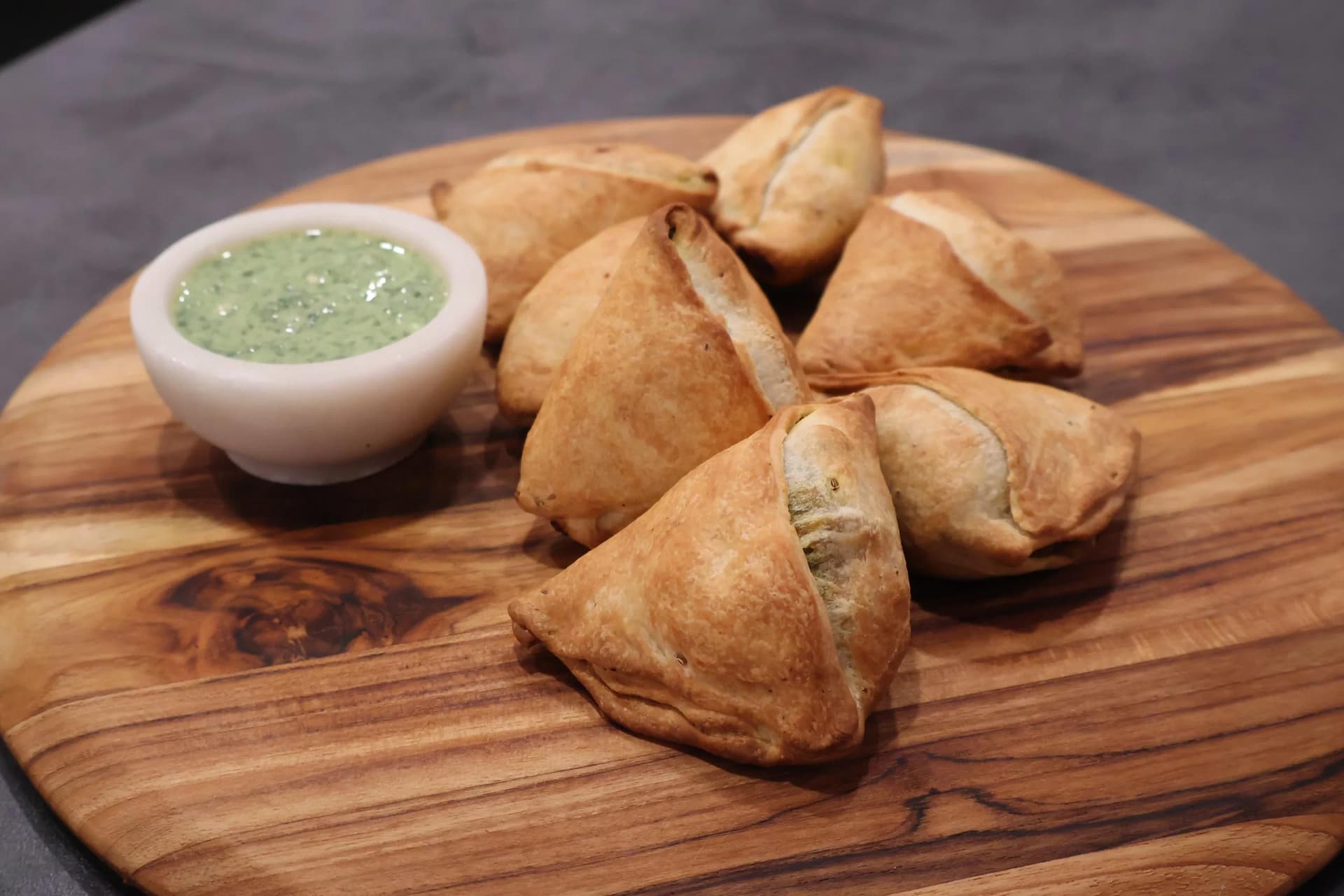 Air fryer potato samosas on wooden board with green cilantro chutney dip in white bowl.