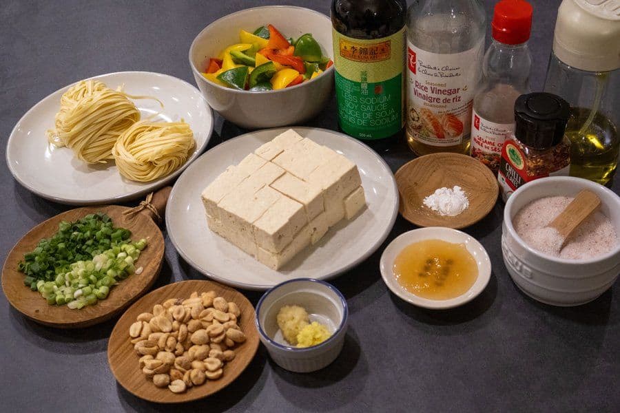 Ingredients for tofu stir fry laid out on a gray surface: noodles, tofu, bell peppers, cashews, green onions, sauces, and spices.