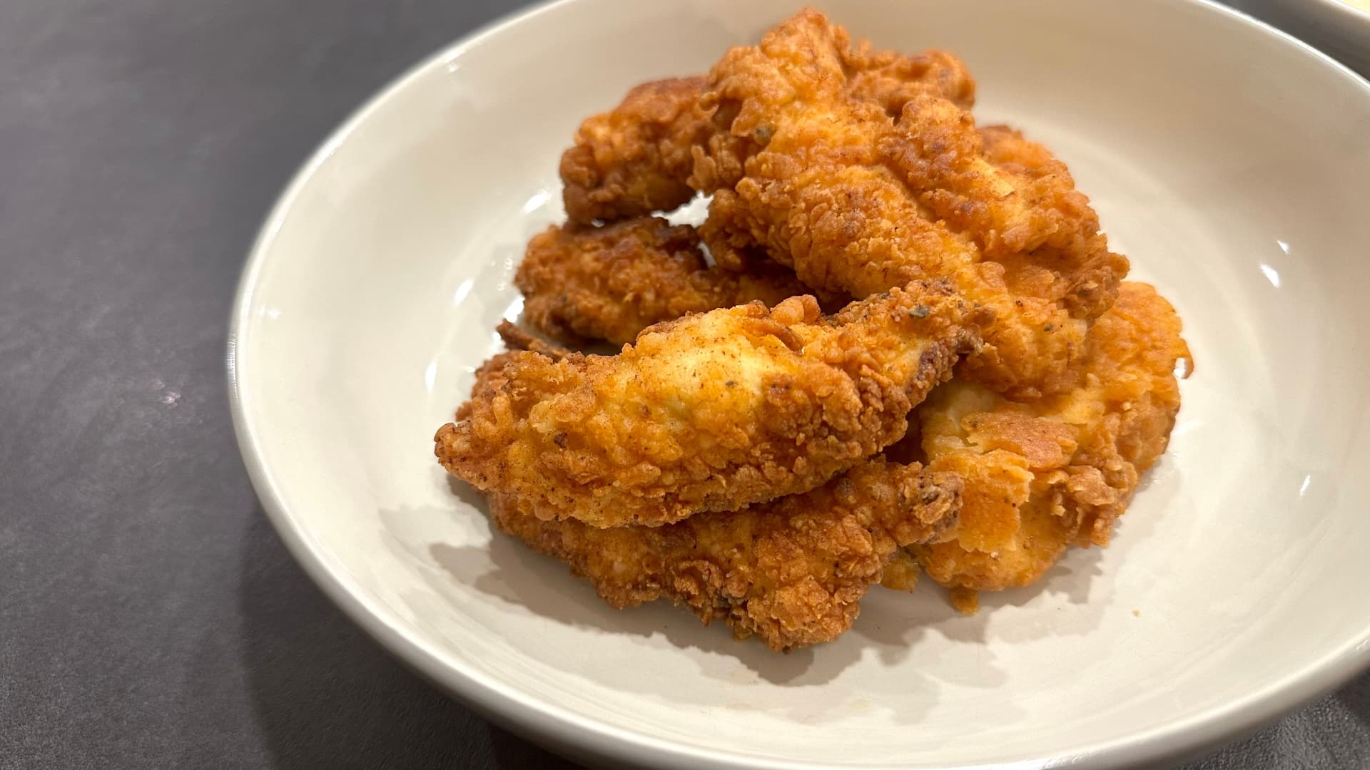 Golden-brown crispy fried chicken tenders stacked on a white plate against a gray surface.