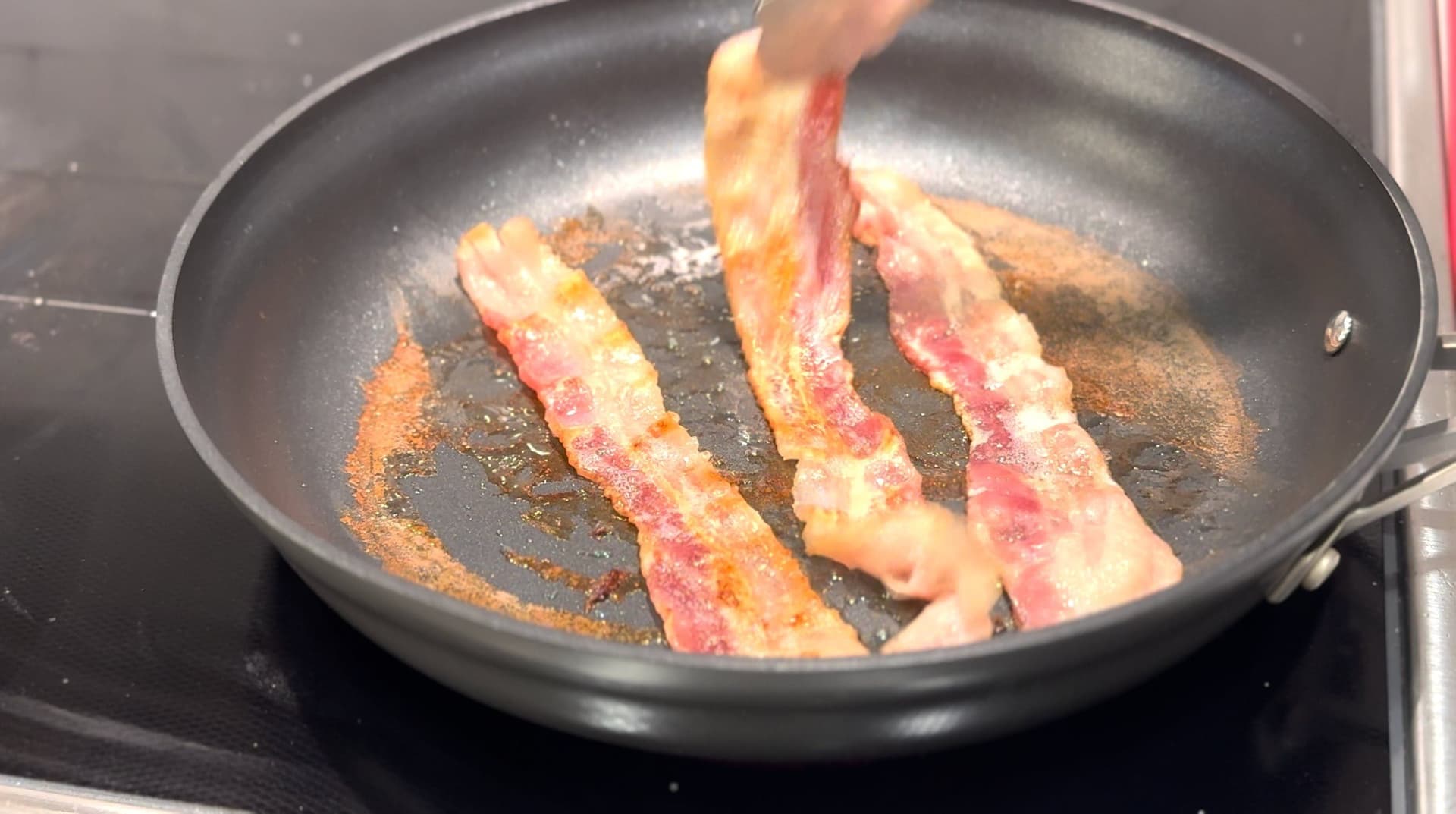 Three strips of bacon cooking in a skillet with water and rendered fat on medium-low heat.