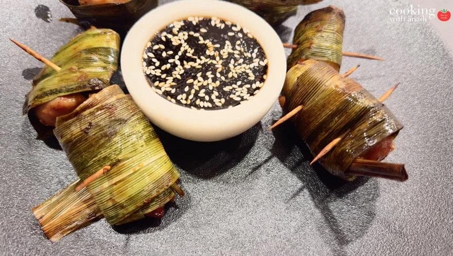 Pandan-wrapped chicken parcels secured with toothpicks, arranged around a dark dipping sauce topped with sesame seeds.