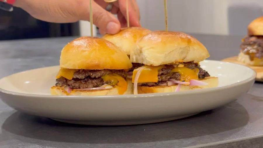 Two smash burger sliders on a white plate with melted cheese, caramelized onions, and what appears to be ham, served on soft dinner rolls.