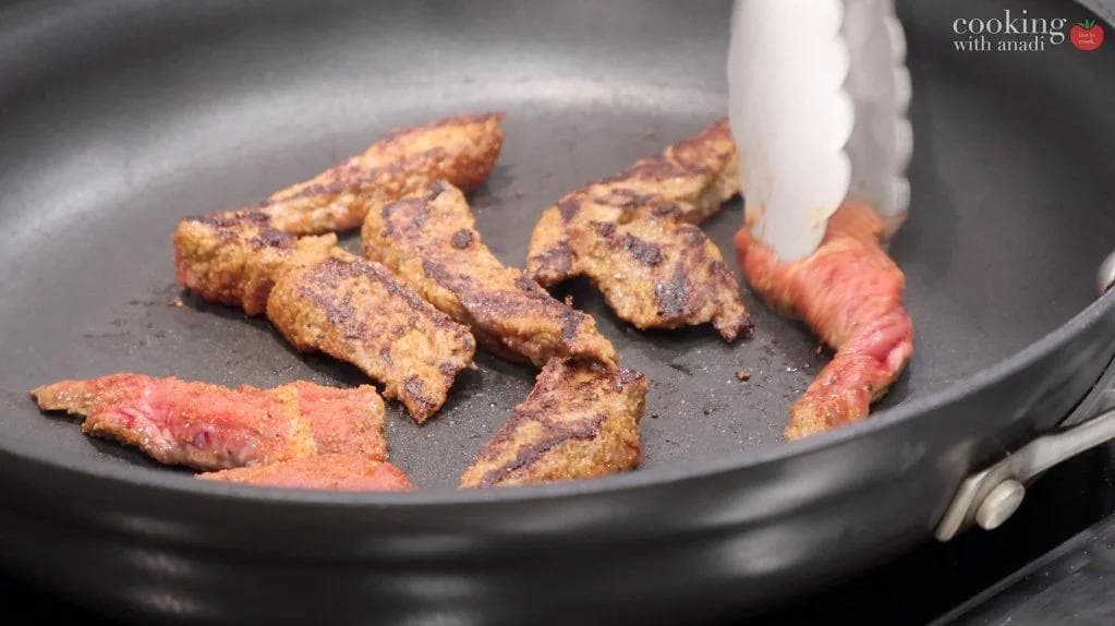 Seasoned steak strips searing in a cast iron skillet over medium heat, showing golden-brown crust.