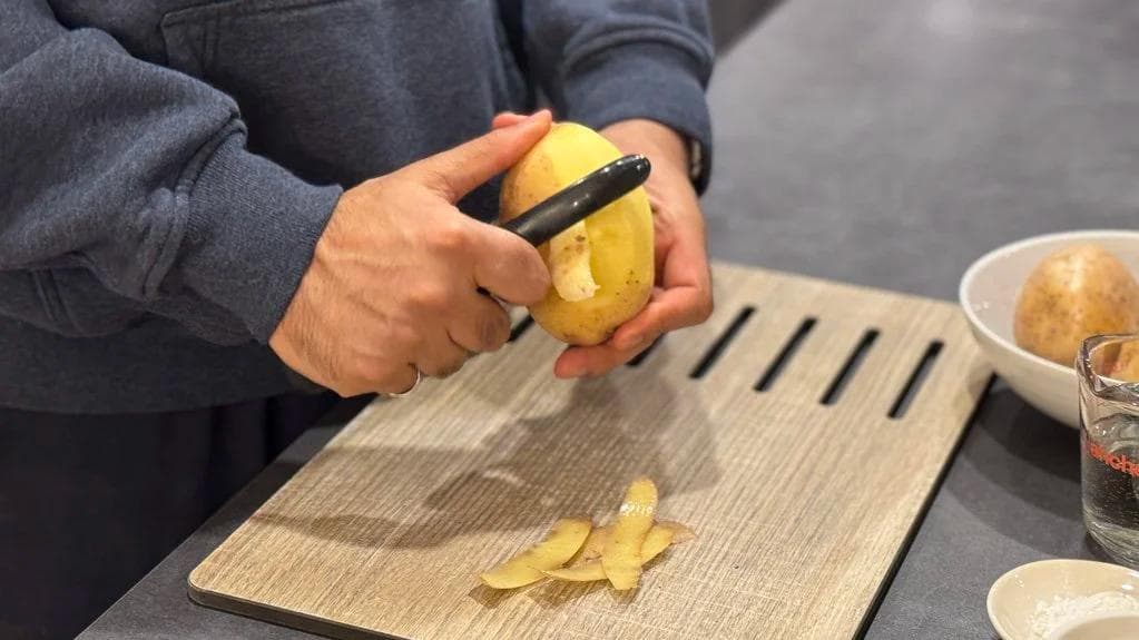 Hands peeling and slicing a yellow potato on a wooden cutting board with a knife.