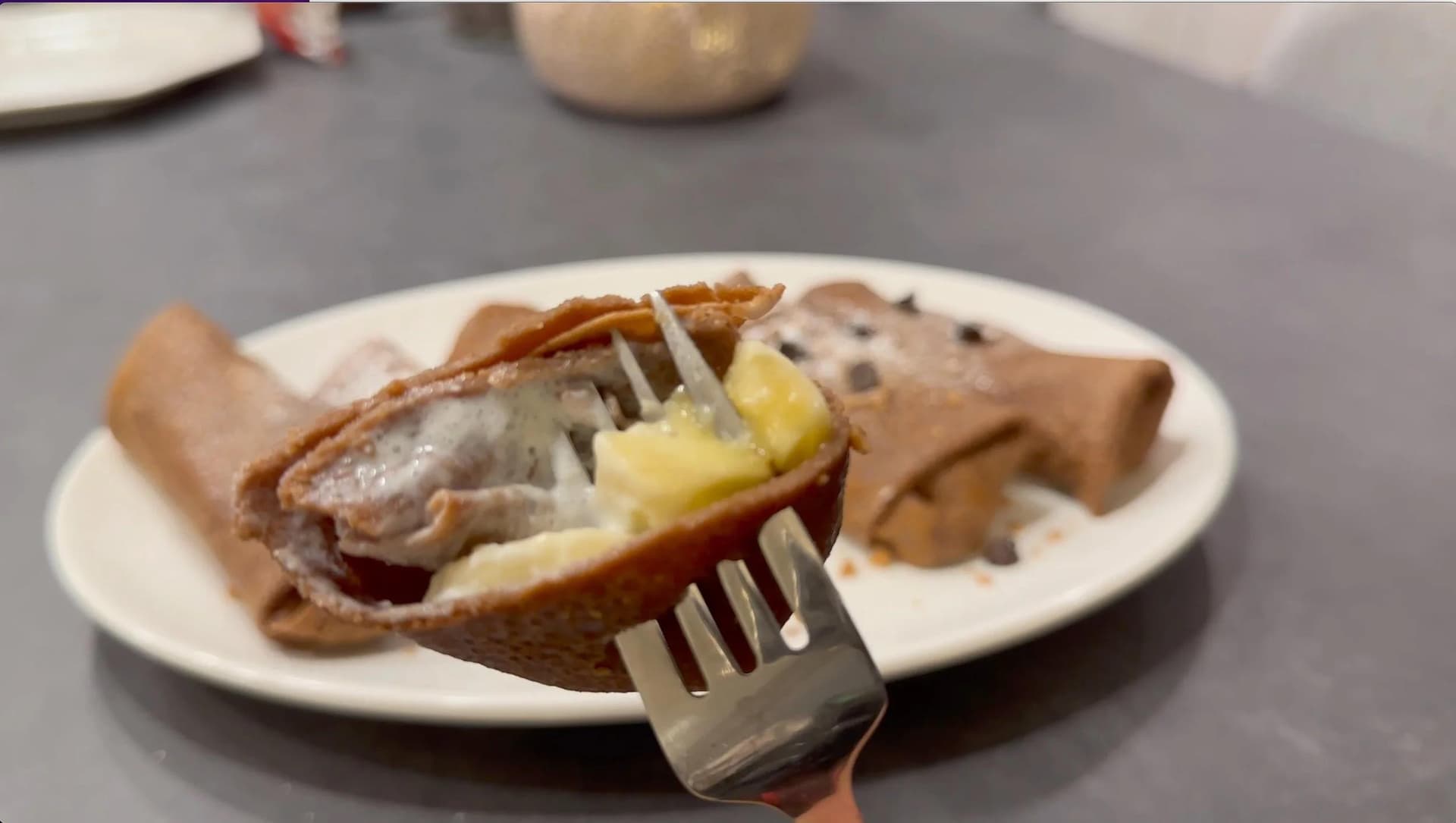 Fork cutting into a chocolate crepe filled with cream and topped with powdered sugar on a white plate.