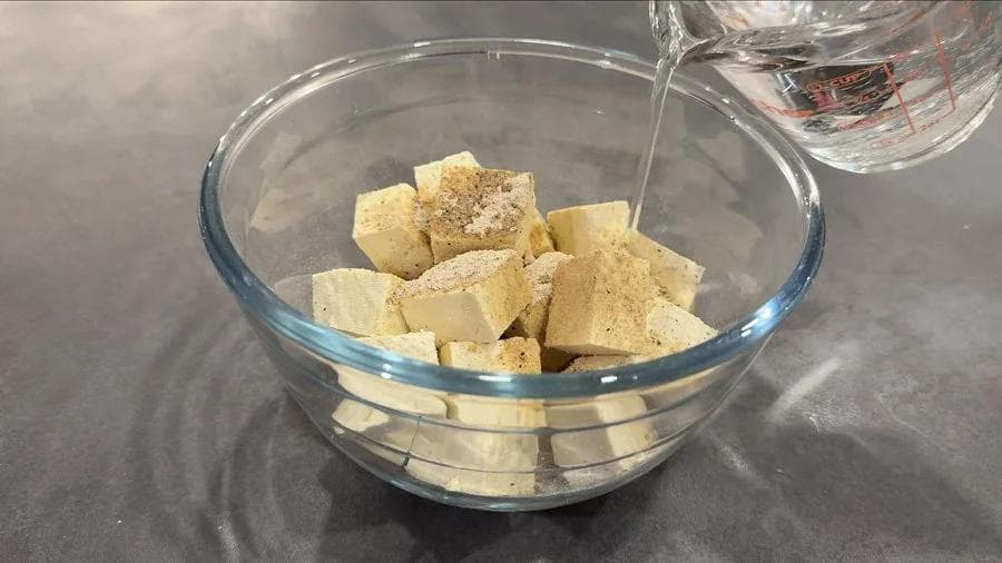 Cubed tofu coated with spices in a glass bowl with water being poured in.