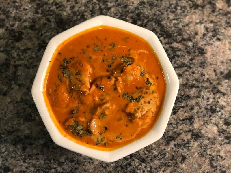 Butter chicken curry in a white bowl, garnished with fresh cilantro, showing a creamy orange sauce with tender chicken pieces.