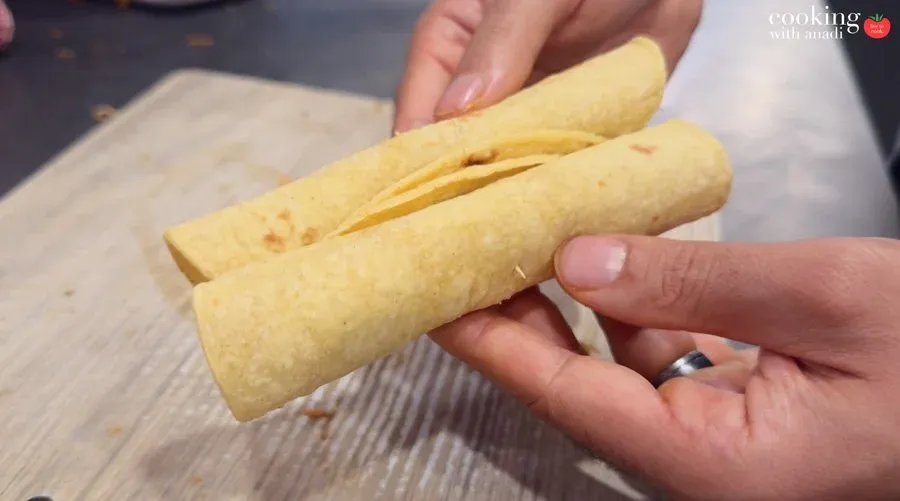 Two tightly rolled corn tortilla taquitos held together with a toothpick, seam-side up, over a wooden cutting board.