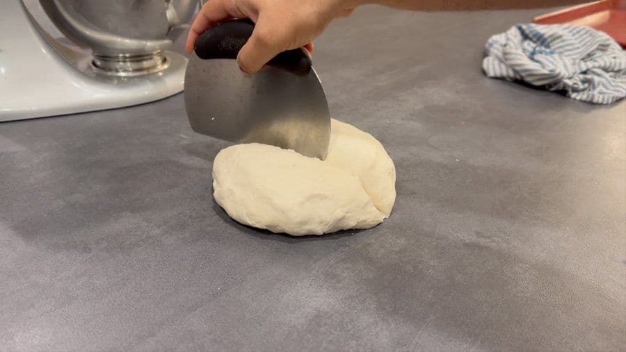 Hand using a bench scraper to fold pizza dough on a gray countertop.