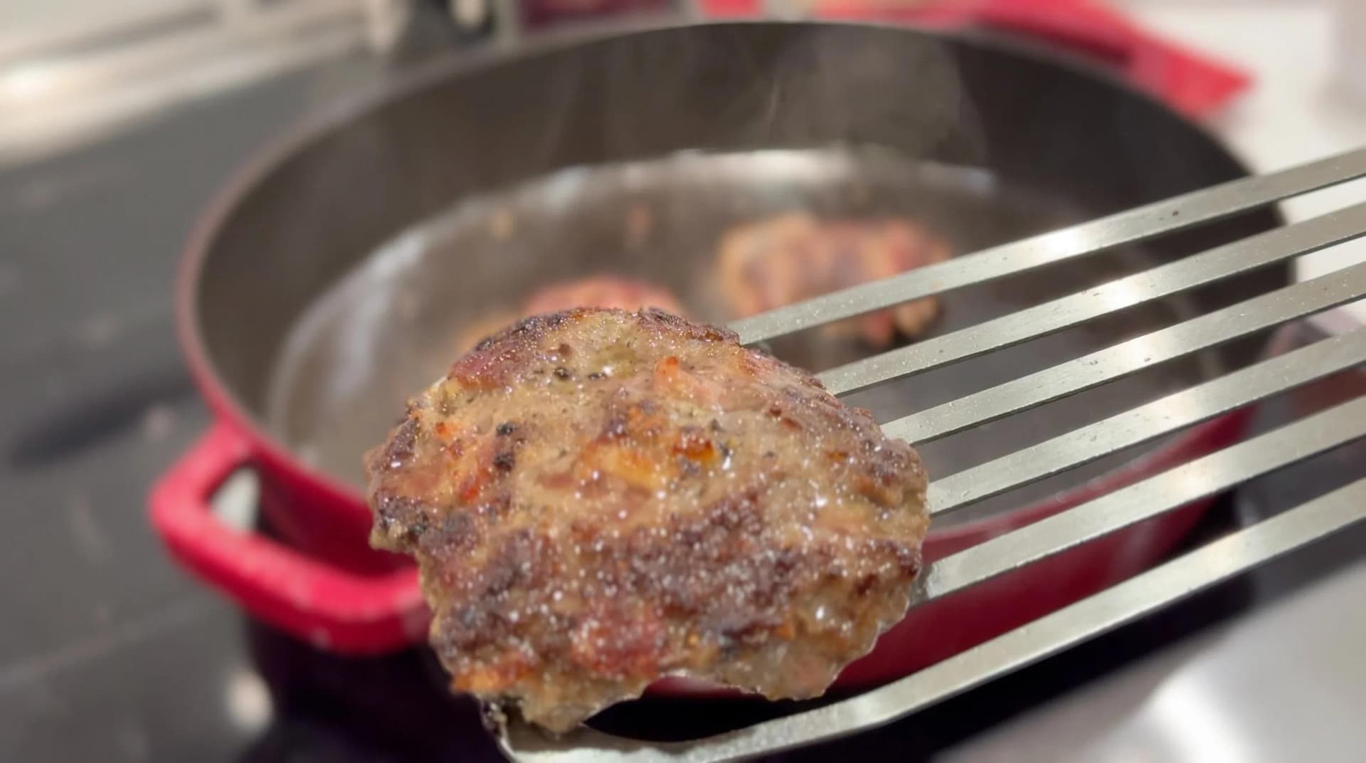 Cooked burger patty being lifted on a spatula from a red cast iron pan, showing golden-brown seared surface.