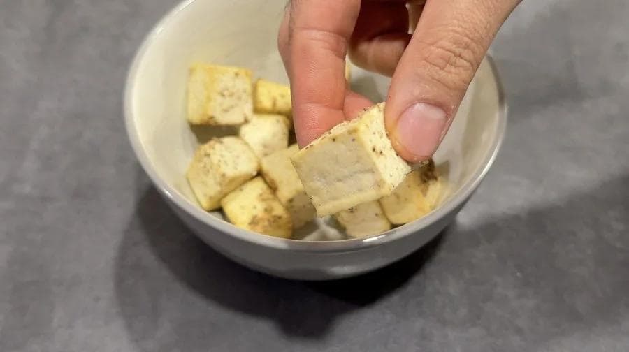 Cubed tofu pieces coated in cornstarch in a white bowl, being picked up with fingers.
