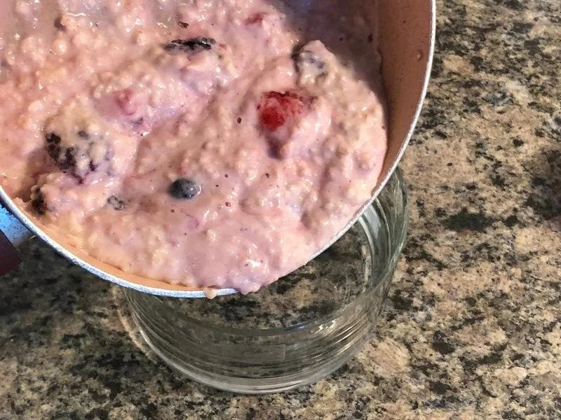 Pink berry oatmeal being poured from a saucepan into a glass bowl, studded with blueberries and raspberries.