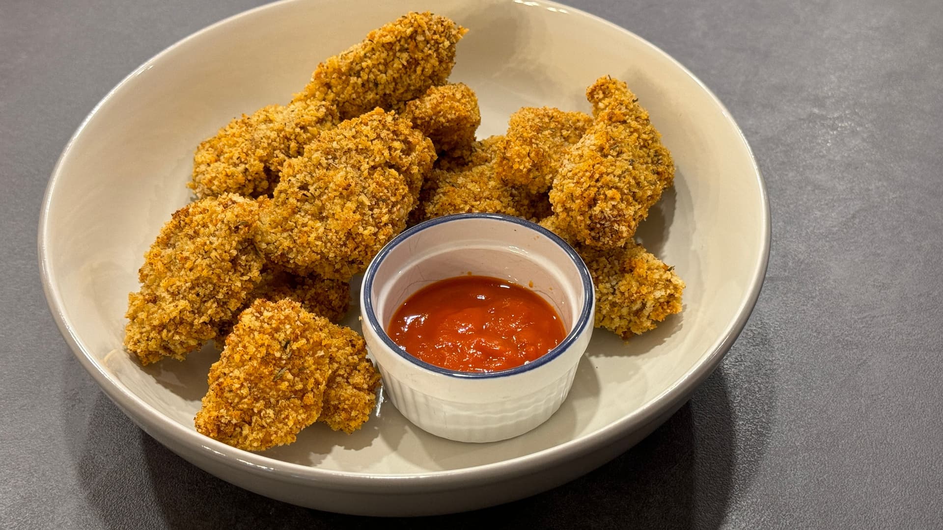 Golden crispy panko chicken nuggets arranged on a white plate with a small bowl of red dipping sauce in the center.