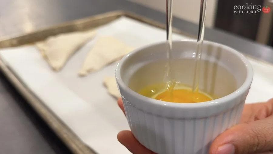 Whisked egg being poured into a white bowl with puff pastry squares visible on a baking sheet in the background.