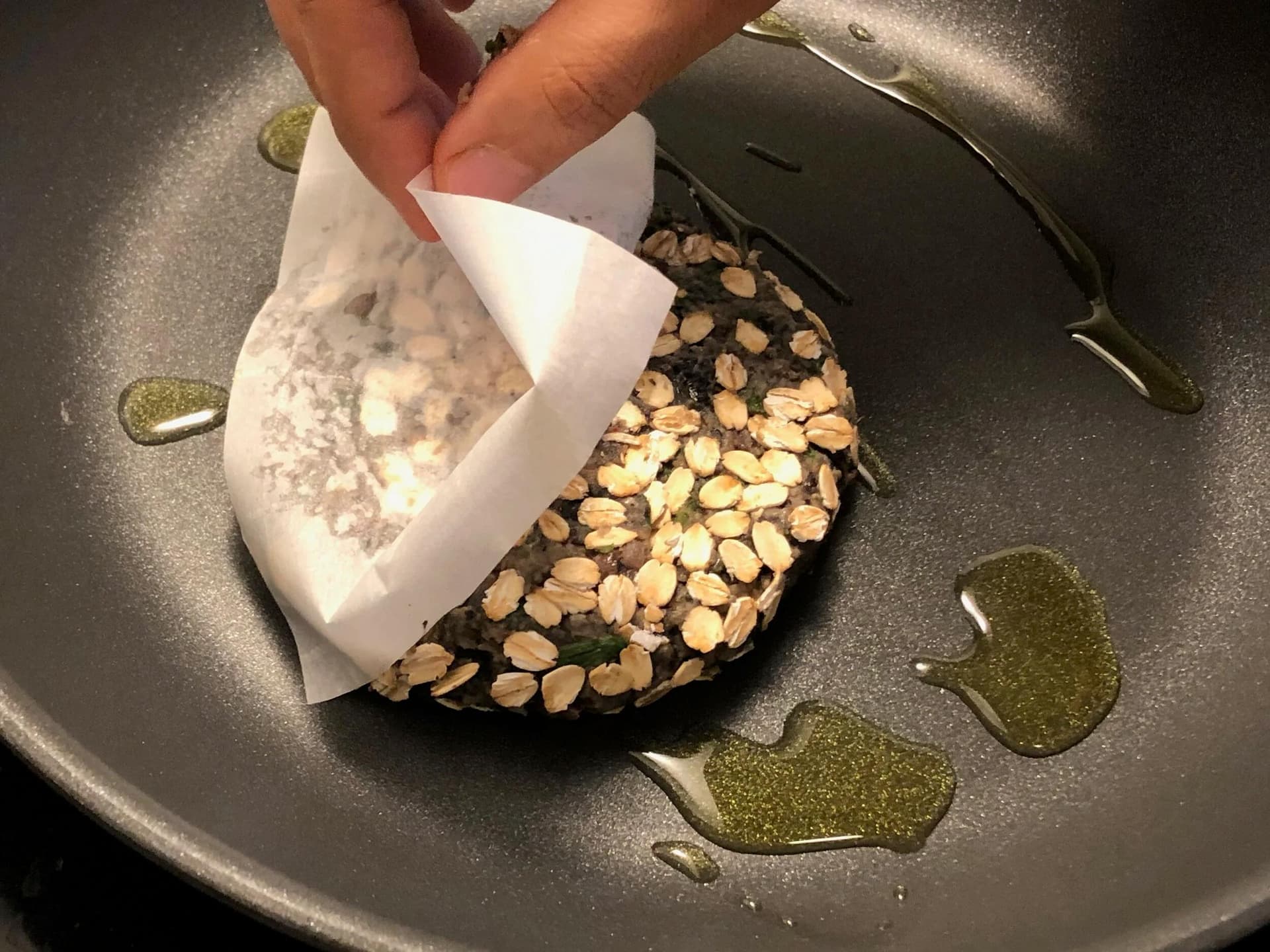 Bean patty with sliced almonds cooking in olive oil in a dark skillet.