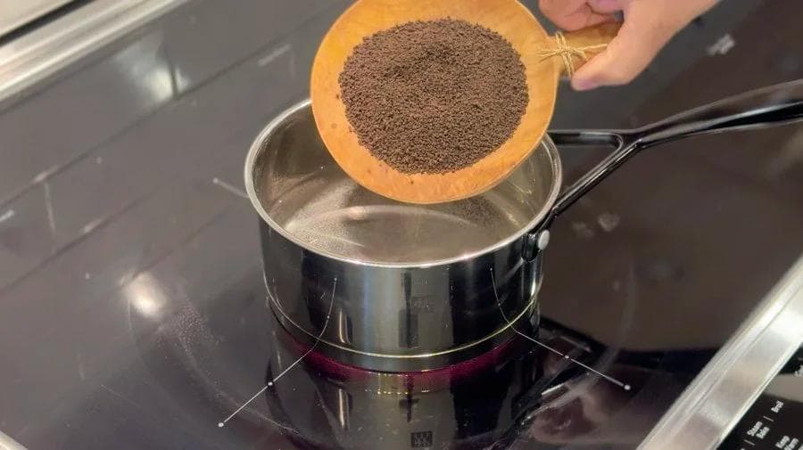 Pouring chai masala spice blend from an orange bowl into a saucepan of simmering water and tea.