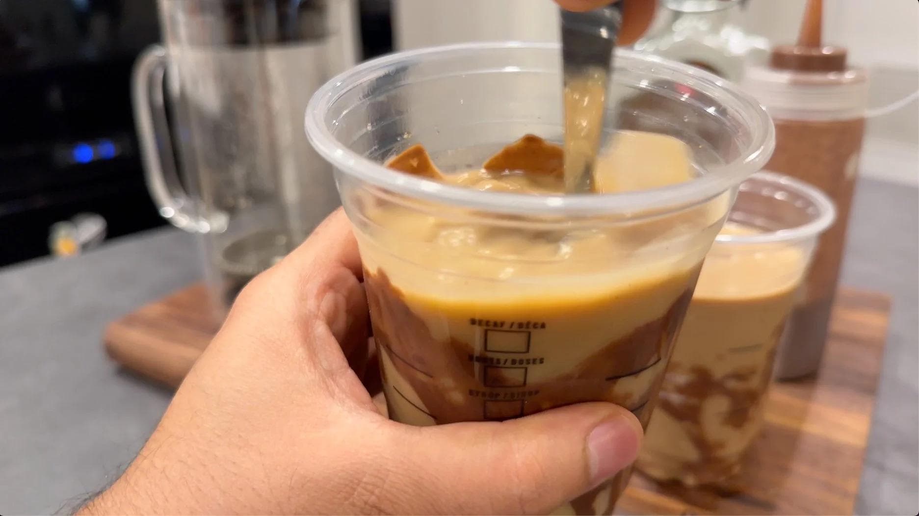 Hand holding a clear cup of iced mocha with chocolate syrup drizzle, ice cubes, and coffee with milk.