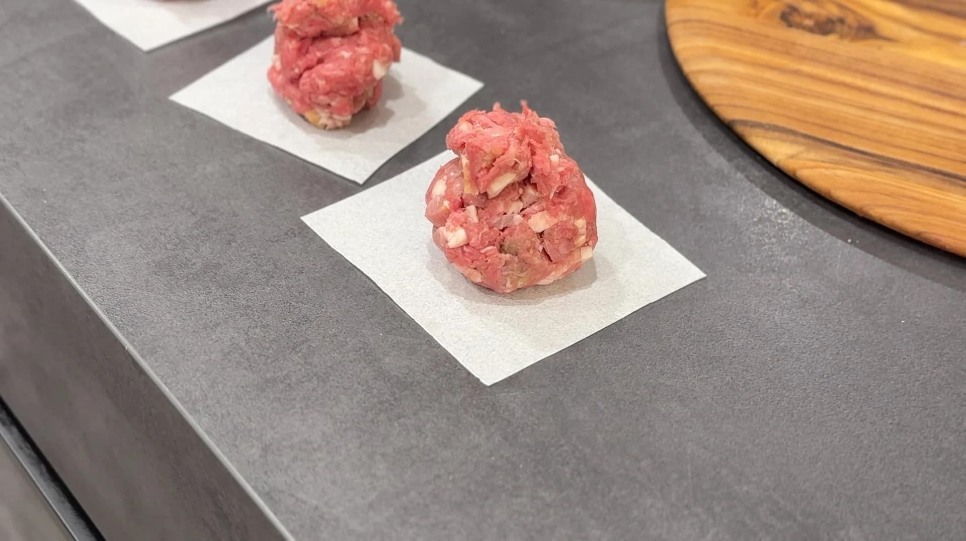 Three raw ground beef patties with bacon and seasonings on parchment paper squares, ready for cooking.