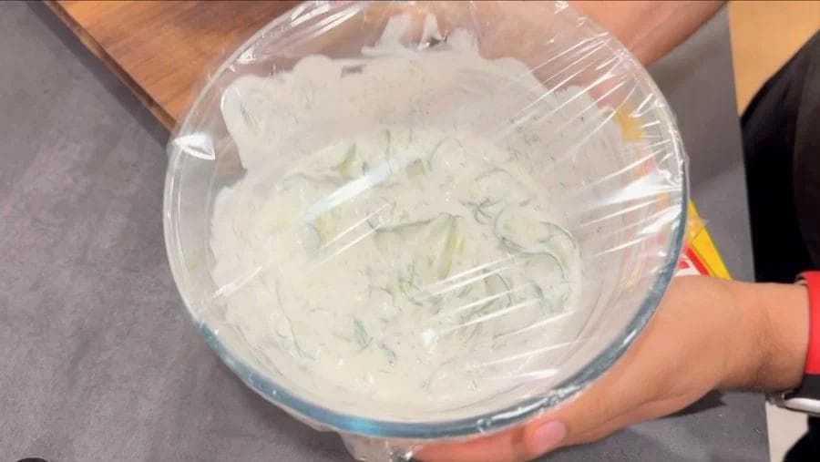 Creamy dill yogurt dressing mixed with cucumber slices in a glass bowl covered with plastic wrap.