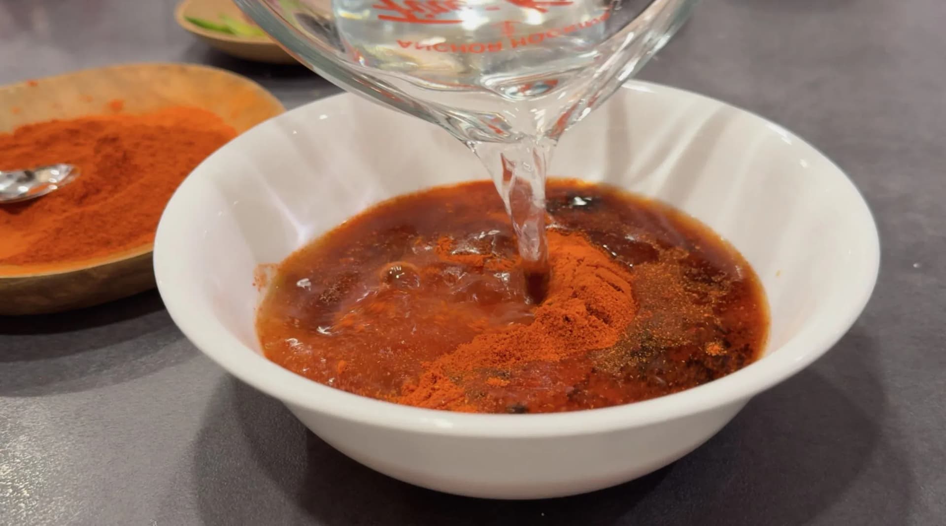 Water being poured from a glass into a white bowl containing red chilli powder, soy sauce, and ketchup sauce mixture.