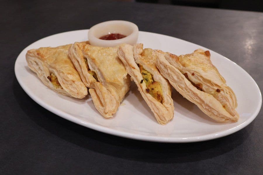 Four golden air-fried aloo patties arranged on a white plate with a small bowl of red sauce.