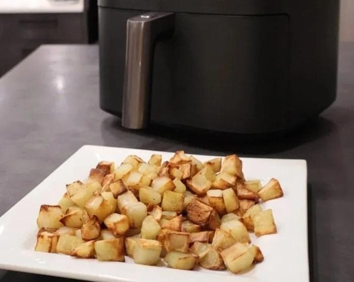 Crispy golden cubed roasted potatoes on a white plate with an air fryer in the background.