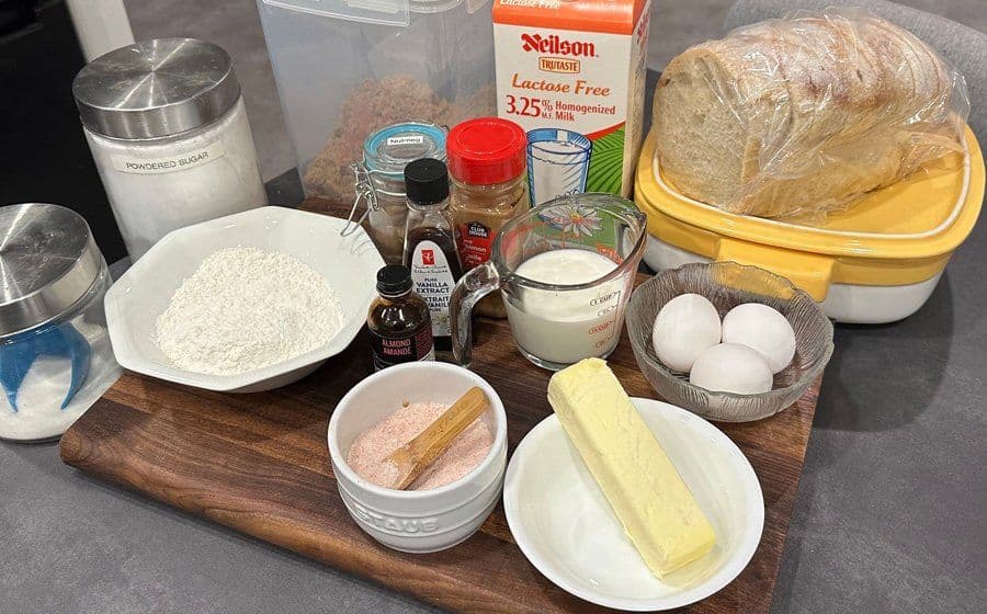 Ingredients for overnight French toast casserole arranged on a wooden board, including eggs, butter, milk, bread, and spices.