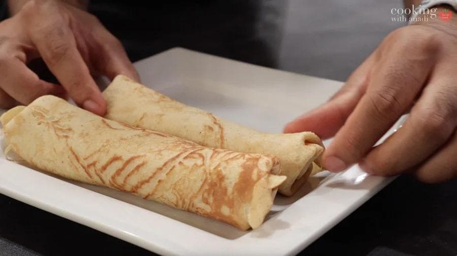 Hands rolling a pumpkin crepe filled with cream cheese into a cigar shape on a white plate.