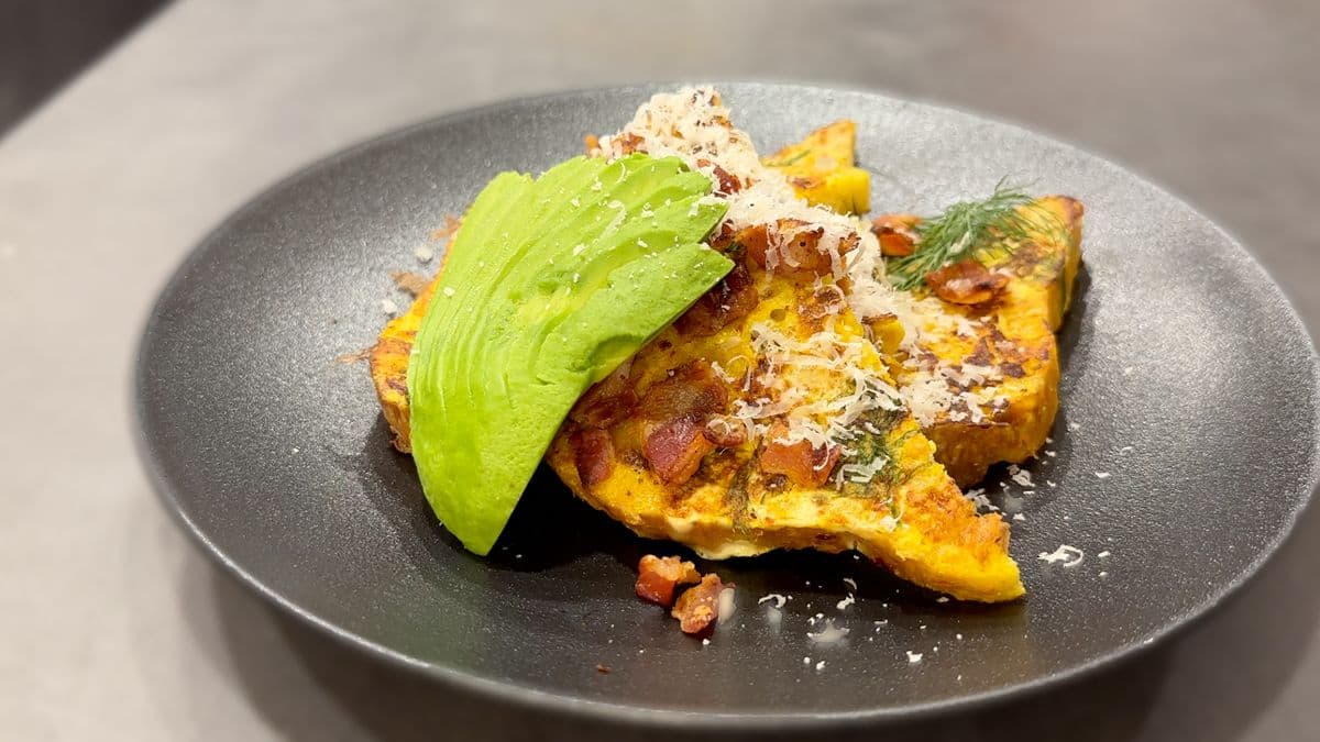 Savory Indian-spiced French toast topped with sliced avocado, crispy bacon, microgreens, and spiced breadcrumbs on a gray plate.