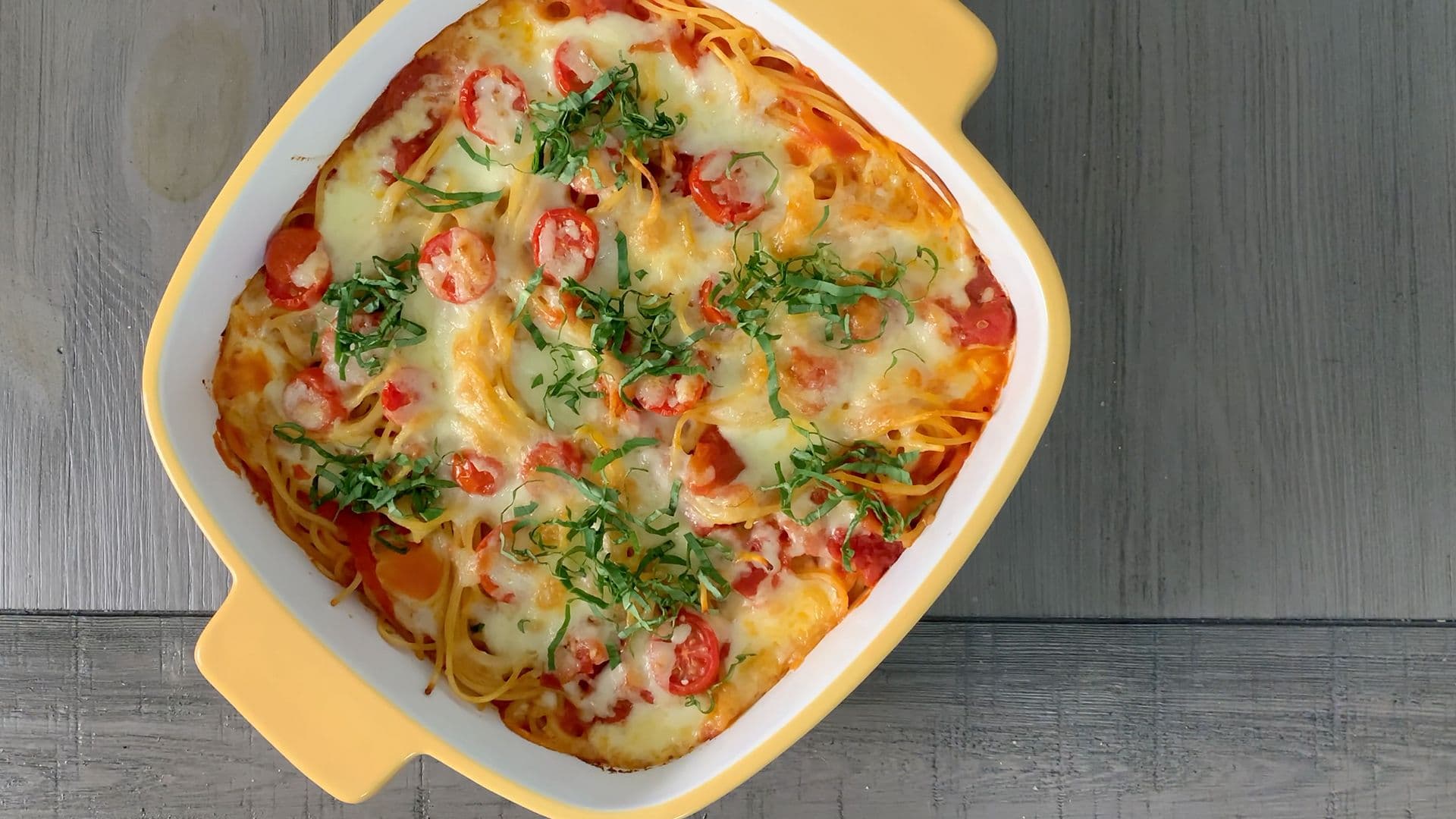 Baked spaghetti casserole in yellow dish topped with melted cheese, tomatoes, and fresh herbs.