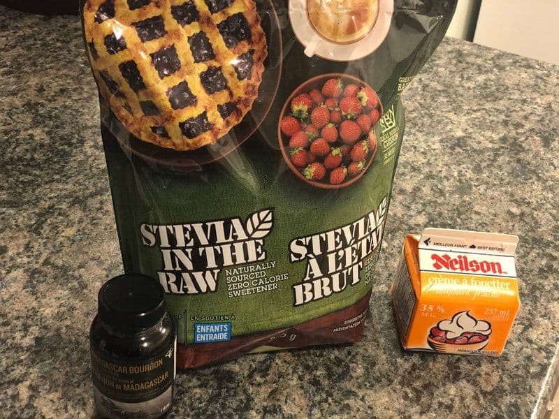 Ingredients for homemade whipped cream displayed on a granite countertop: Stevia sweetener packets, Madagascar bourbon vanilla bean paste, Neilson whipped cream, and fresh raspberries.