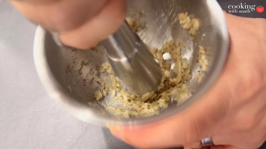 Crushing garlic and white peppercorns into a paste using a metal pestle in a small stainless steel mortar.